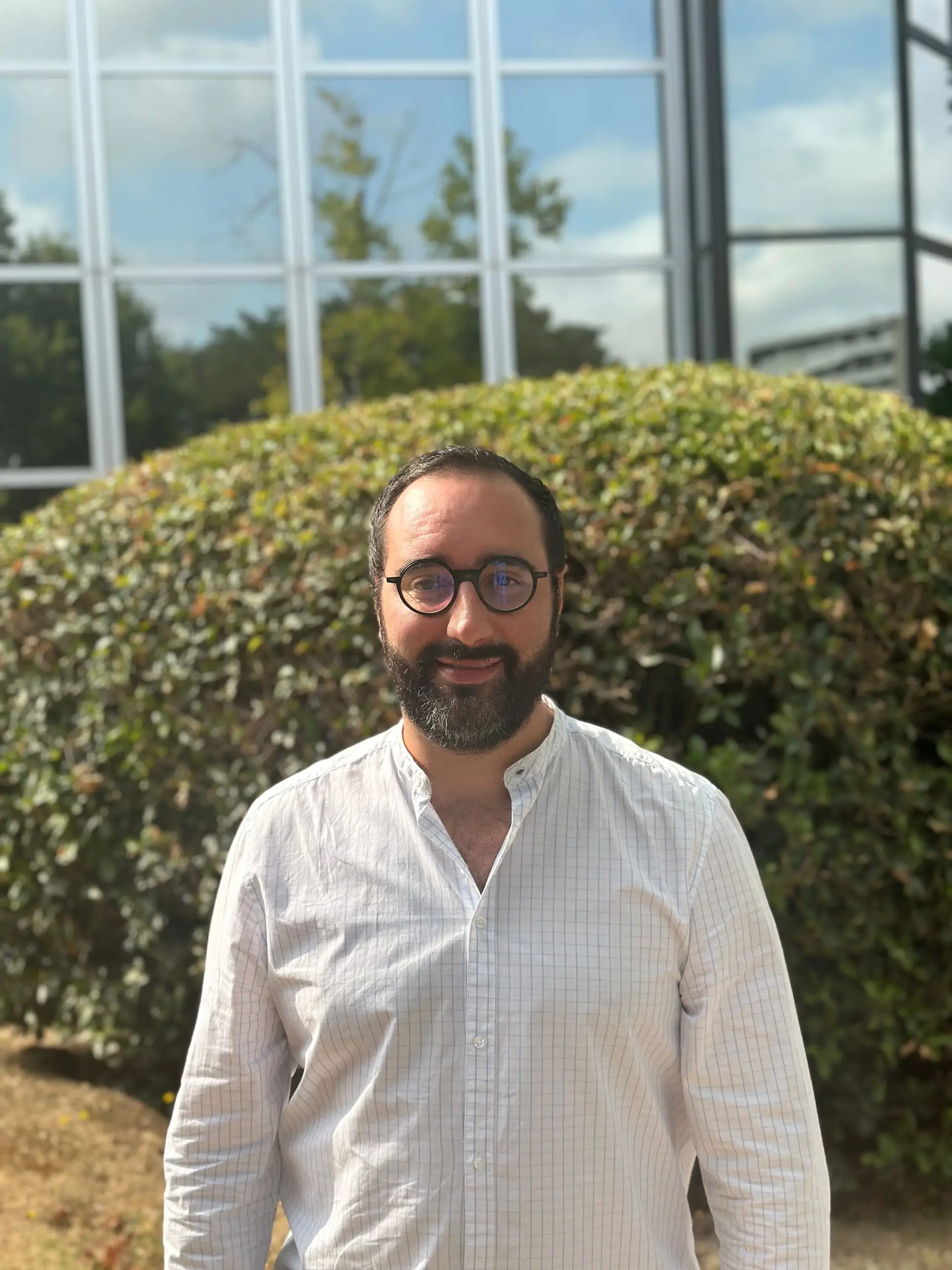 Portrait de Clément Larrieu avec une barbe et des lunettes rondes devant une haie et une façade vitrée.