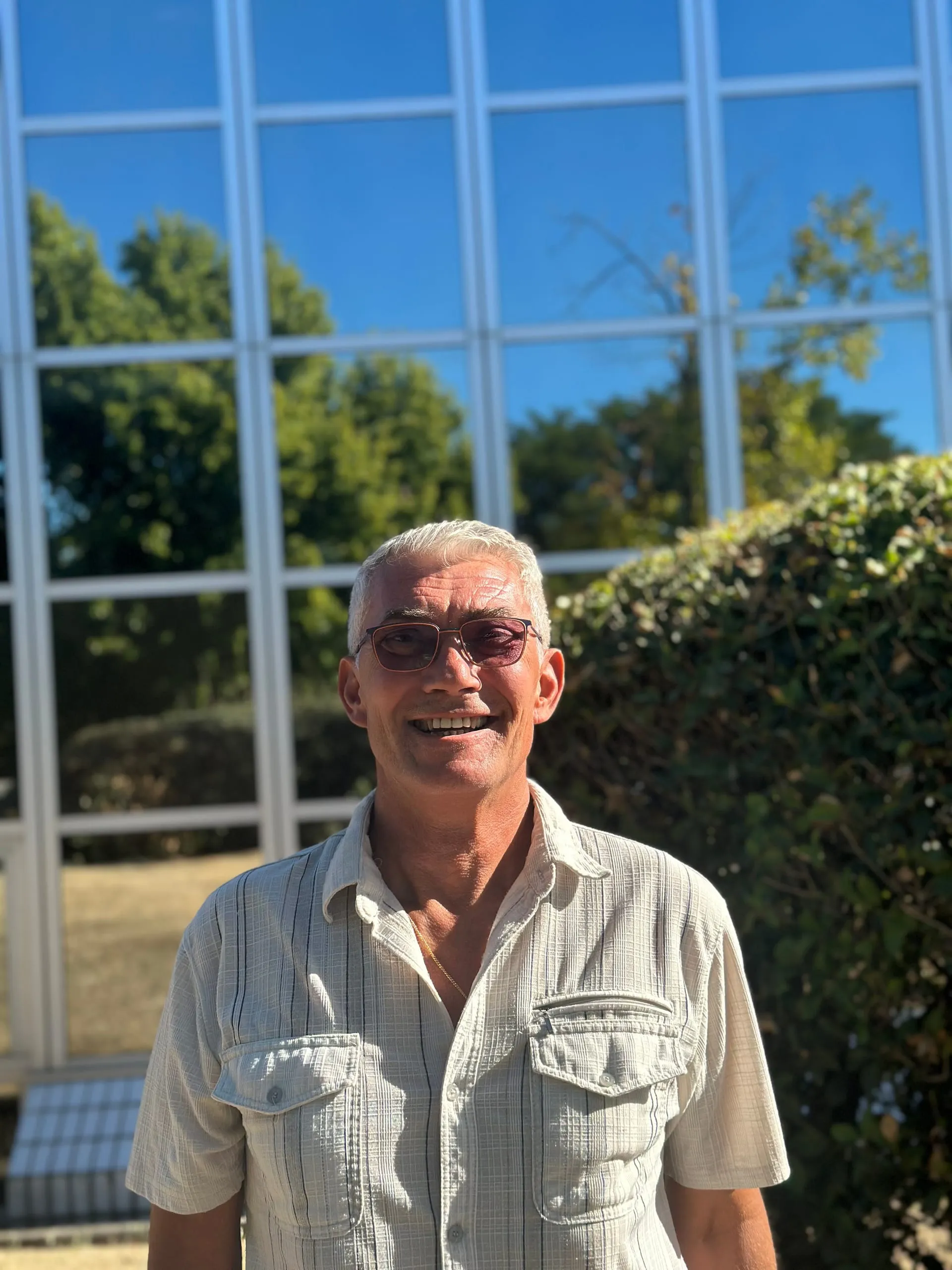 Portrait de Yann Hamon souriant, avec des lunettes et une chemise claire devant une façade vitrée
